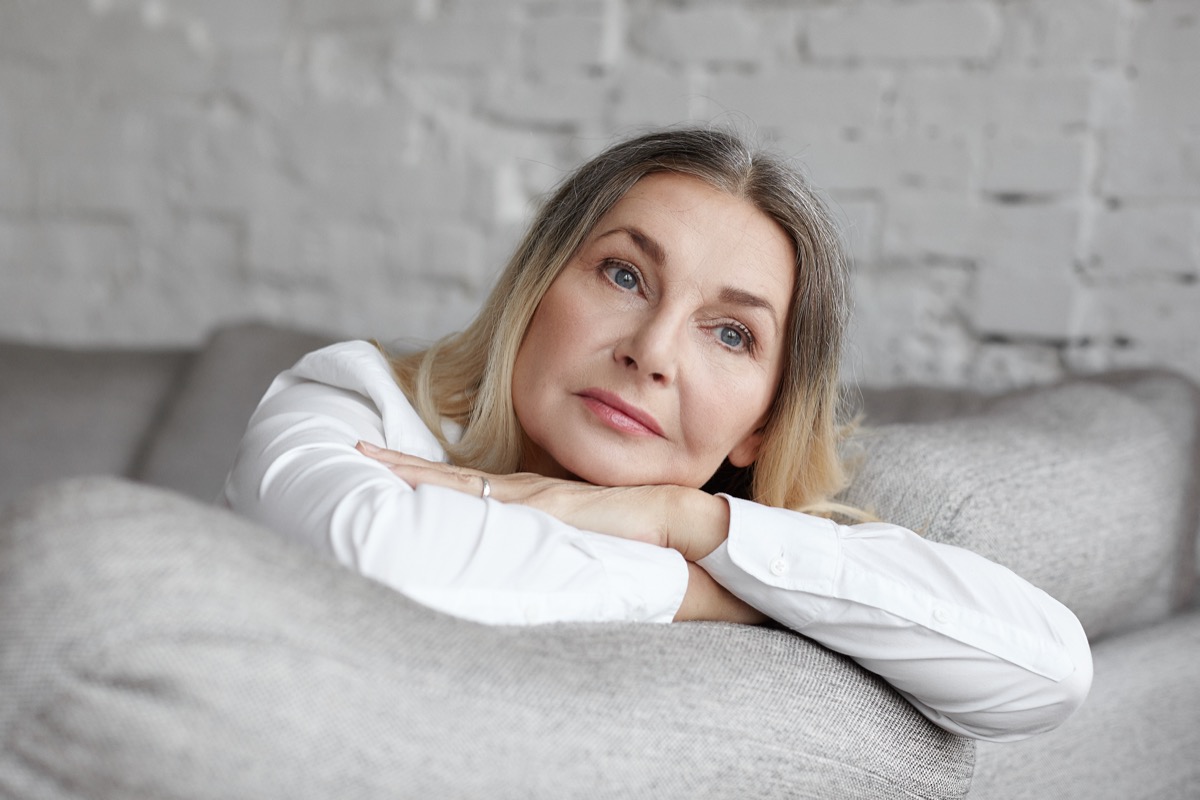 Middle aged woman with long straight hair resting on grey comfortable sofa, having sad unhappy expression.