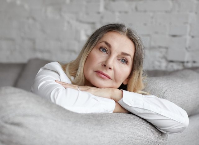 Middle aged woman with long straight hair resting on grey comfortable sofa, having sad unhappy expression.