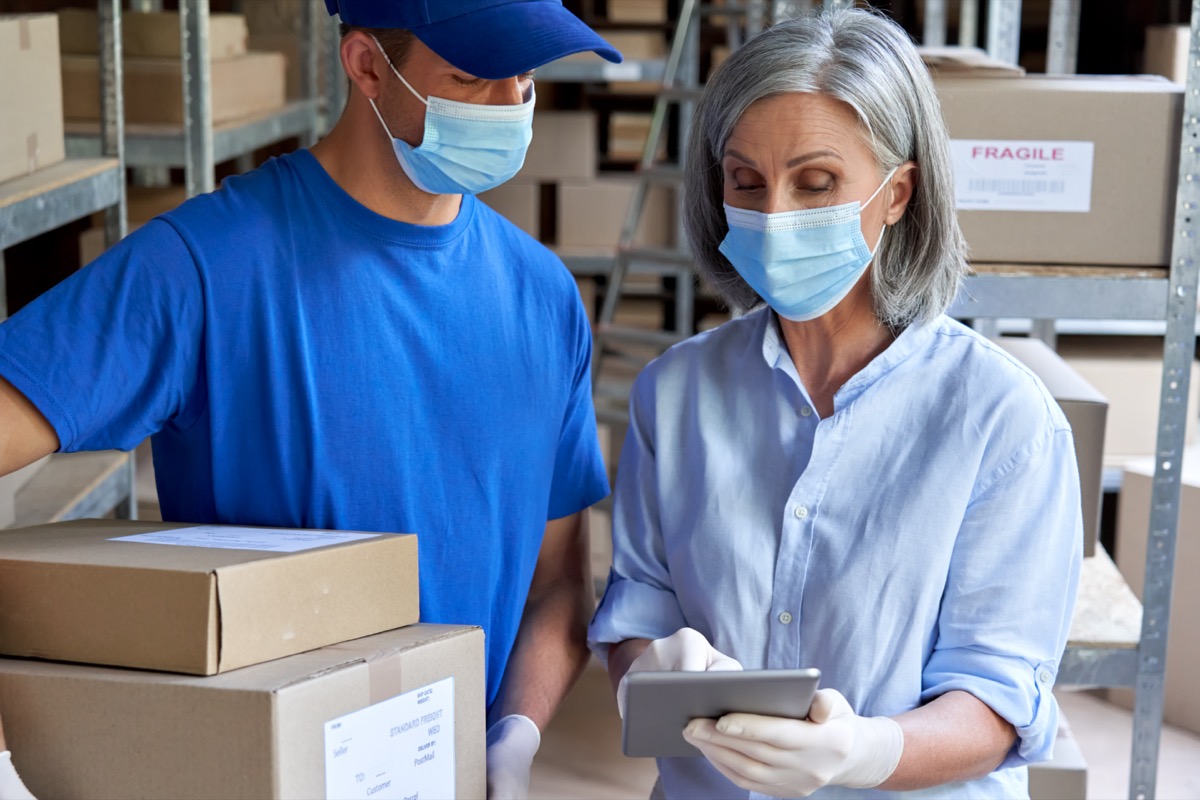 Female supervisor wearing face mask using digital tablet in warehouse talking to male courier holding shipping parcels boxes delivering packages.