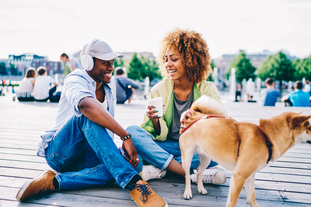 Cheerful afro american male and female hipster joking and having fun sitting on square with cute dog, romantic couple in love laughing during first date on sunny spring day talking to each other