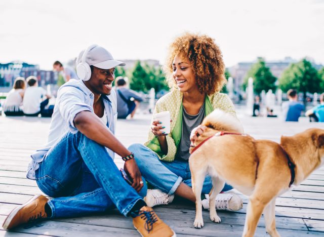 Cheerful afro american male and female hipster joking and having fun sitting on square with cute dog, romantic couple in love laughing during first date on sunny spring day talking to each other