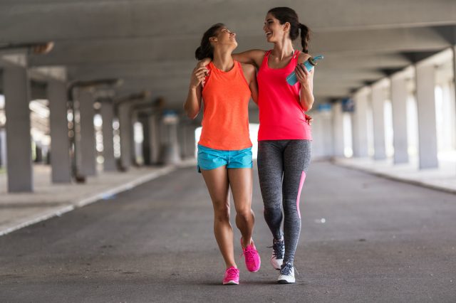 A couple of female friends run on a city street under a city road overpass.  They are relaxing and having fun after running.  Hug each other.  walkers