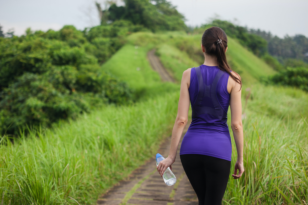 Cô gái mảnh mai trong bộ đồ thể thao bó sát với chai nước đứng ngoài trời giữa thiên nhiên mùa hè cỏ xanh, nhìn từ chính giữa.