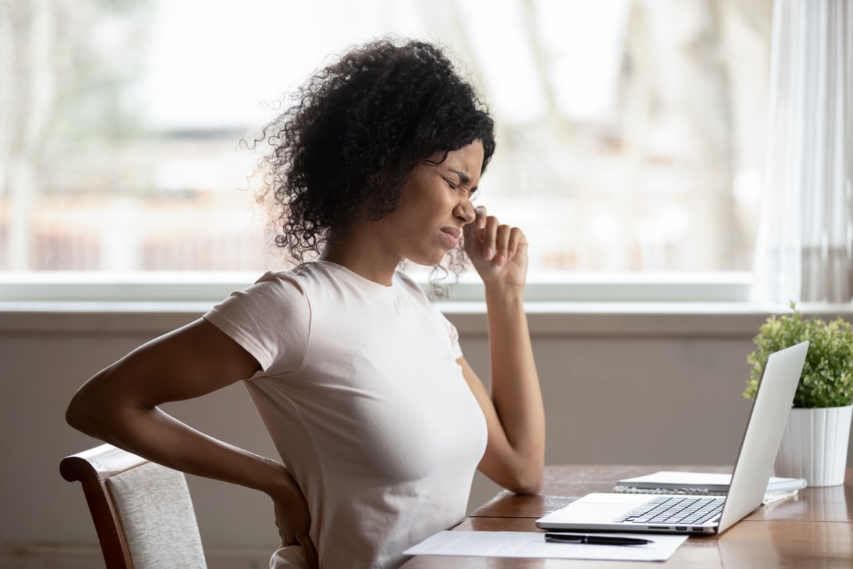 Female worker sit at desk touch back suffer from lower spinal spasm, hurt unhealthy biracial woman stretch have strong backache.