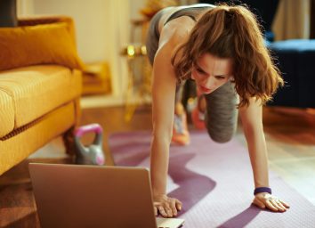 young sports woman in fitness clothes at modern home using online streaming fitness site in laptop and doing circuit training on fitness mat.