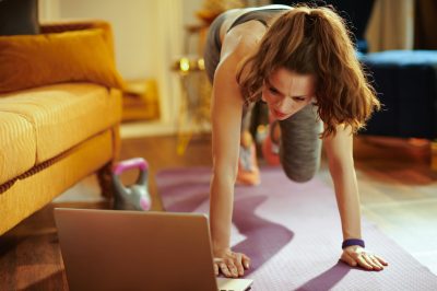 young sports woman in fitness clothes at modern home using online streaming fitness site in laptop and doing circuit training on fitness mat.