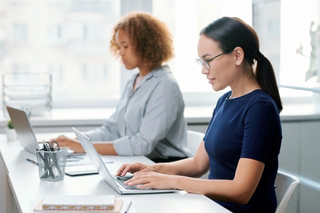 Two women sitting by desk in front of laptops.