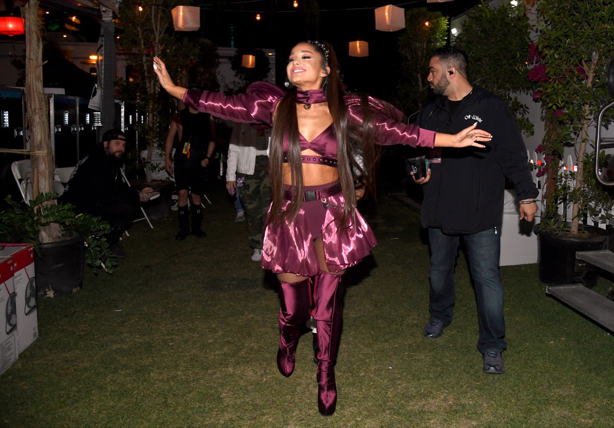 ariana grande smiling with arms outstretched in purple skirt, crop top, and boots