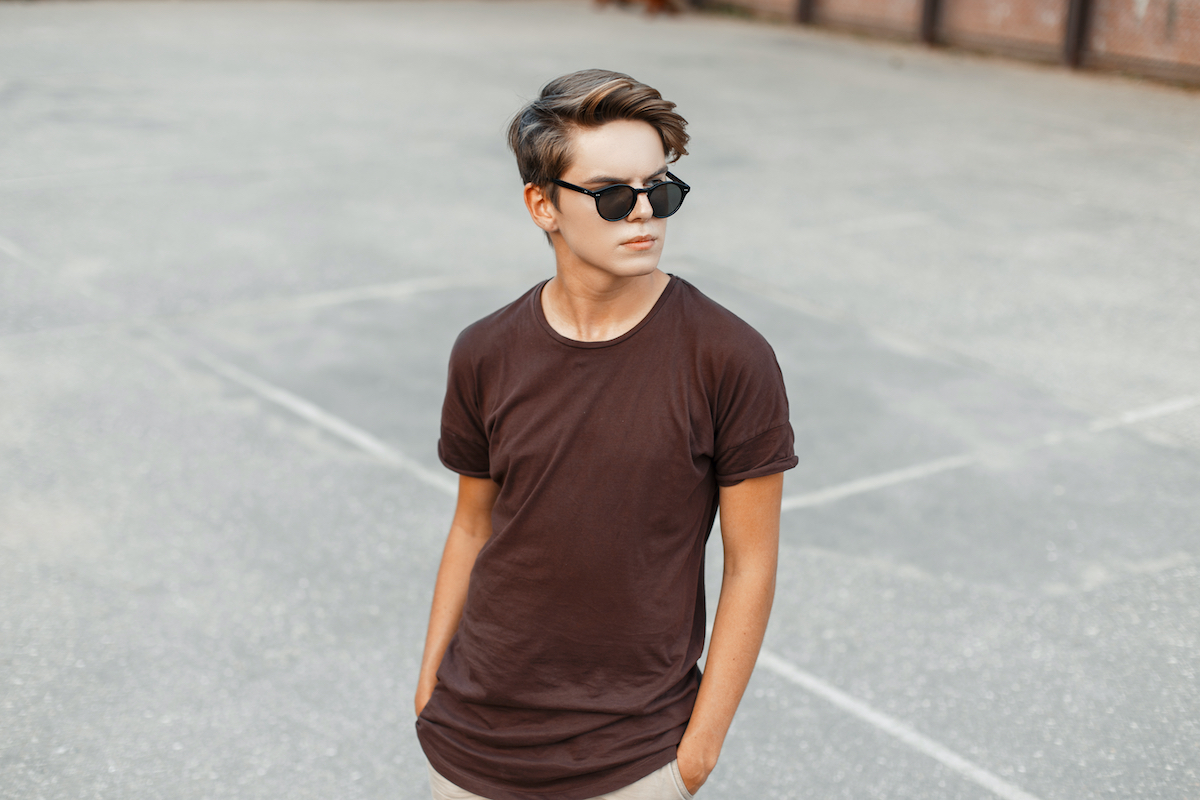 Attractive stylish young hipster man in a brown trendy t-shirt with a stylish hairstyle in fashionable sunglasses on the basketball court. American guy teenage for a walk.