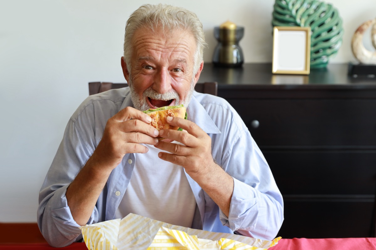 Elderly man eating hamburger in living room with smiling face