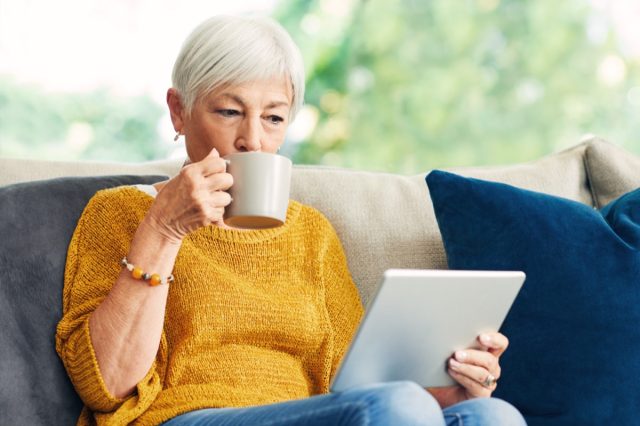 Senior woman using a digital tablet and having coffee on the sofa at home.