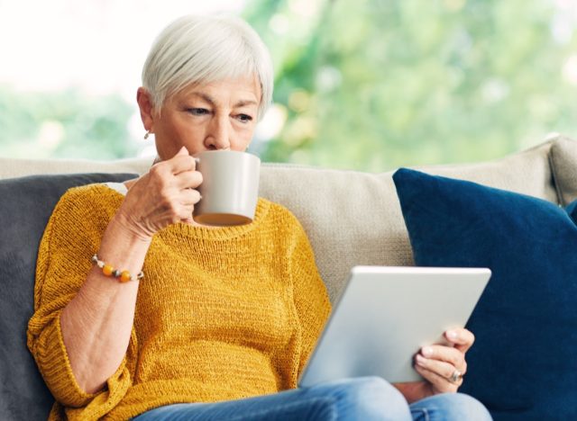 Senior woman using a digital tablet and having coffee on the sofa at home.