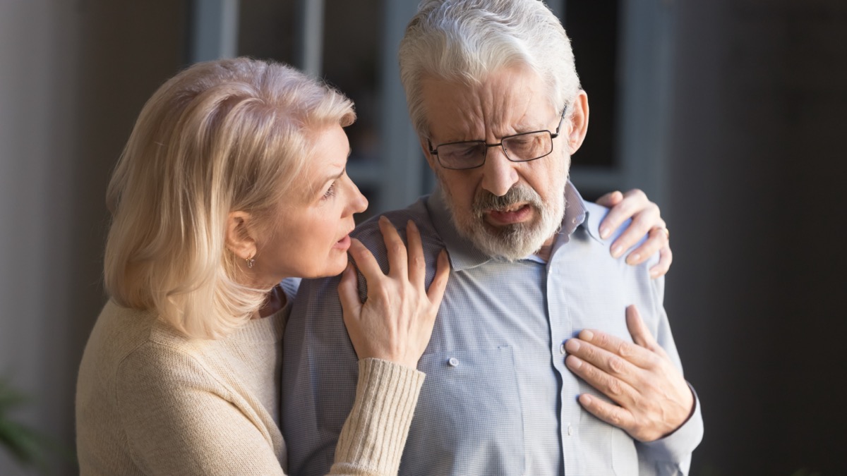 Grey haired man touching chest, feeling pain at home, mature woman supporting him.