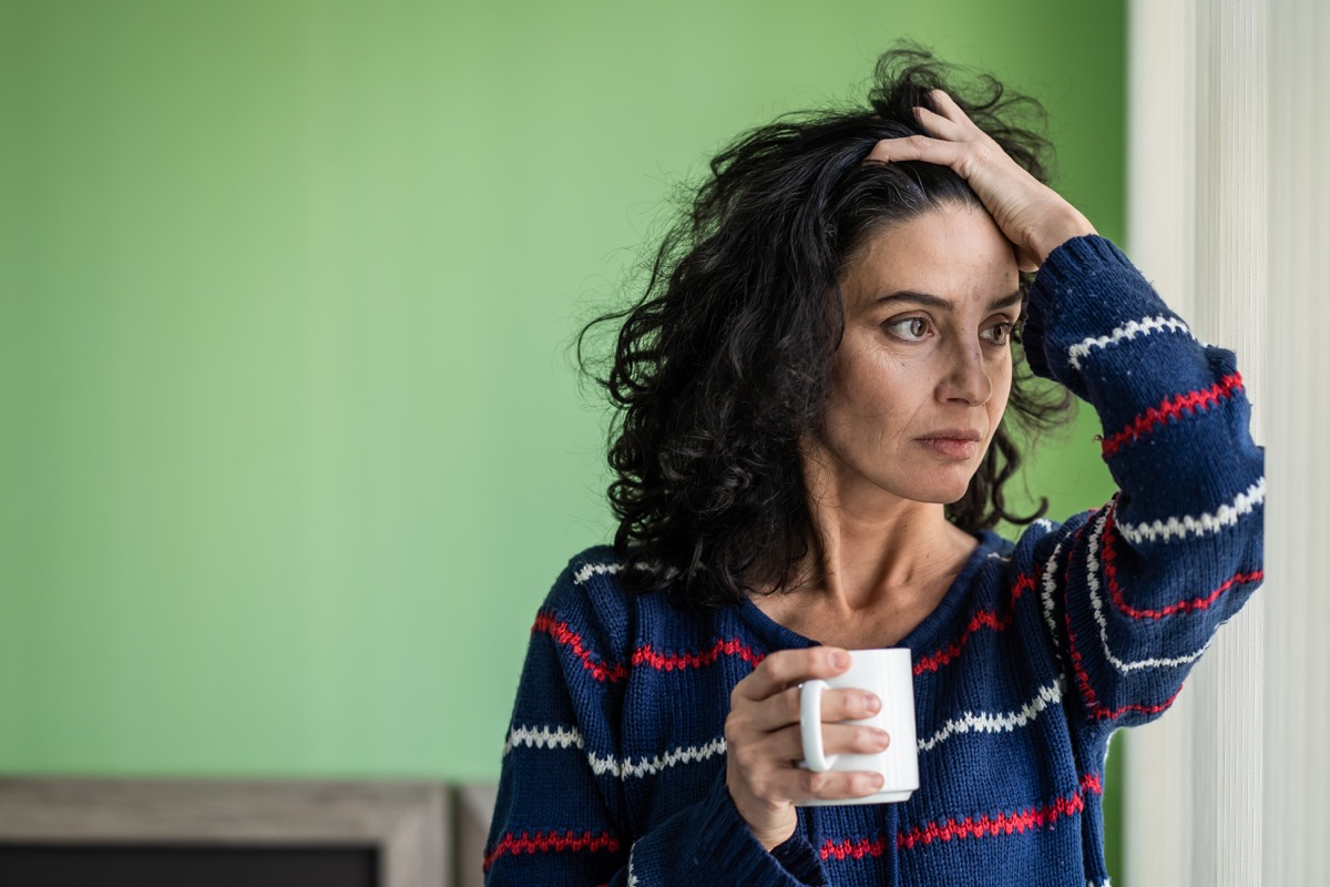 Mature woman standing home alone, worried, drinking coffee.