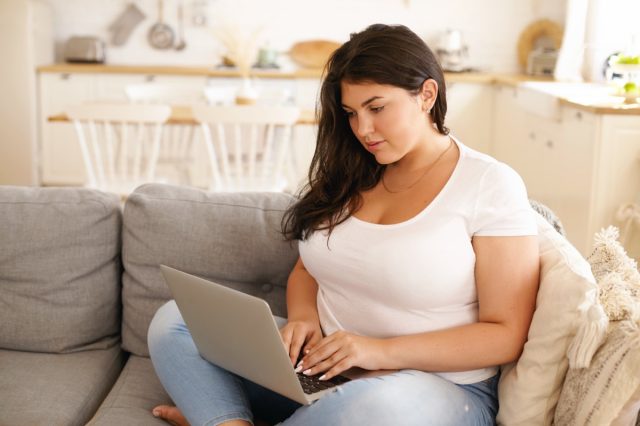 Overweight woman is working on her laptop on a sofa.
