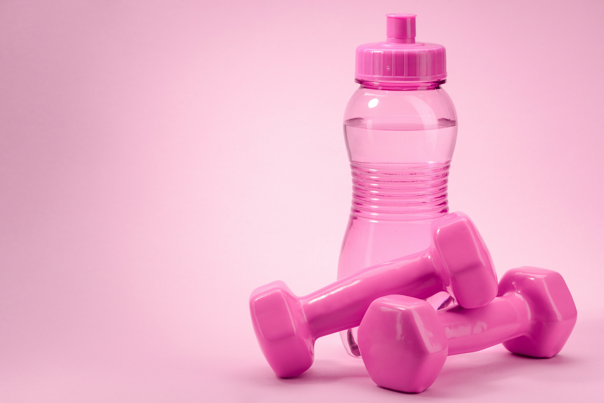 Pink bottle and dumbbellls on pink background with copy space. Gym accessory and equipment of the same color. Fitness and healthy lifestyle.