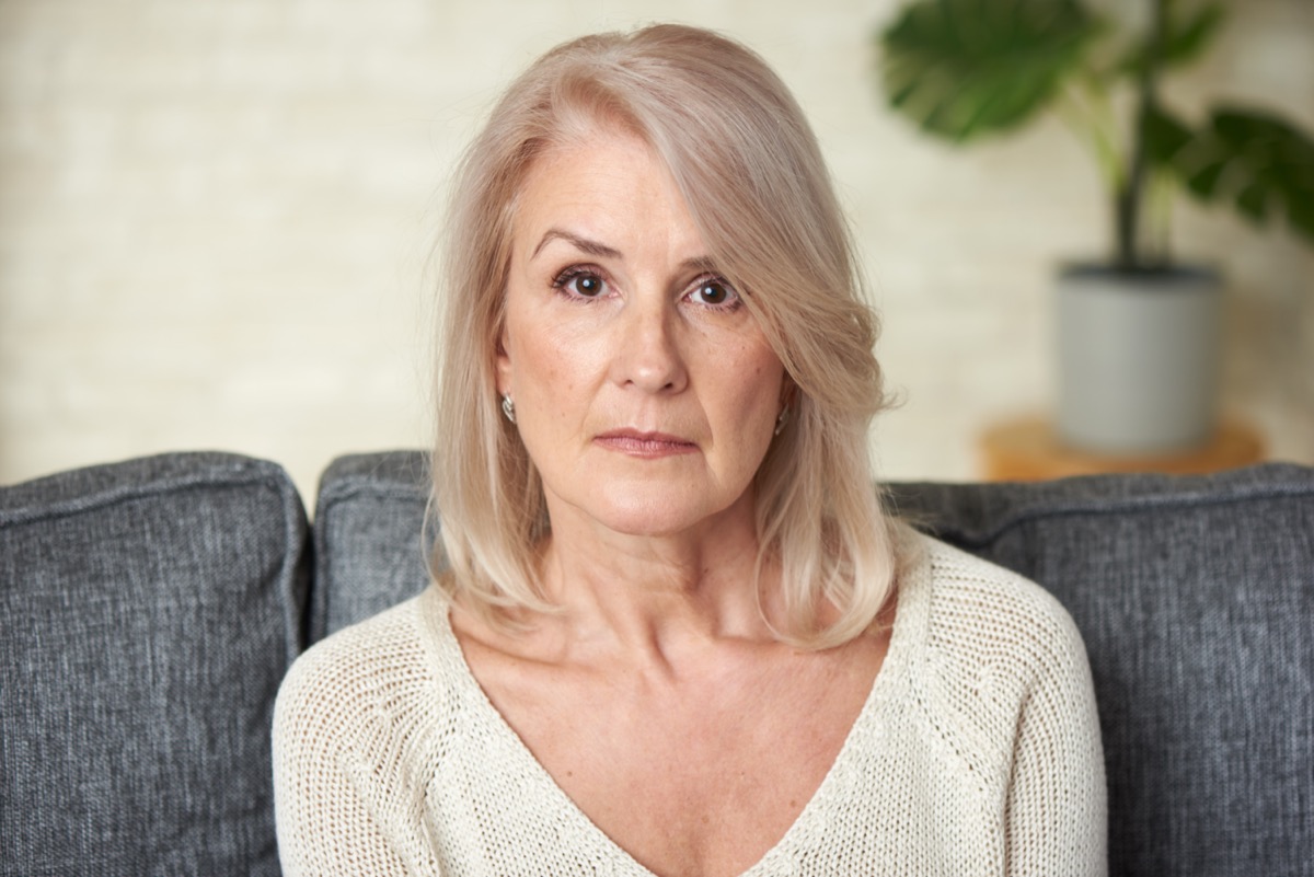 Sad senior blonde woman is sitting on a sofa at home.