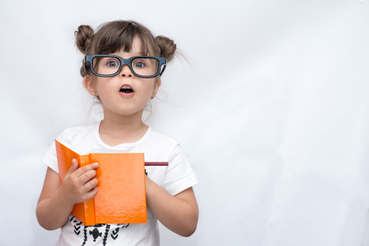 Child wearing glasses, writing in notebook using pencil, keeping mouth wide open.