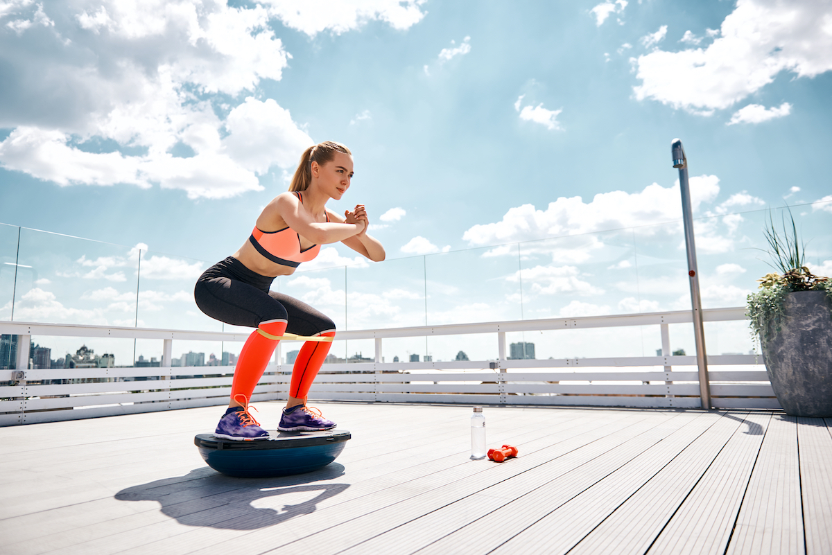 Athlete girl is enjoying work out with outfit on high balcony. She is doing squats on bosu platform while stretching resistance band under knees. Copy space in right side