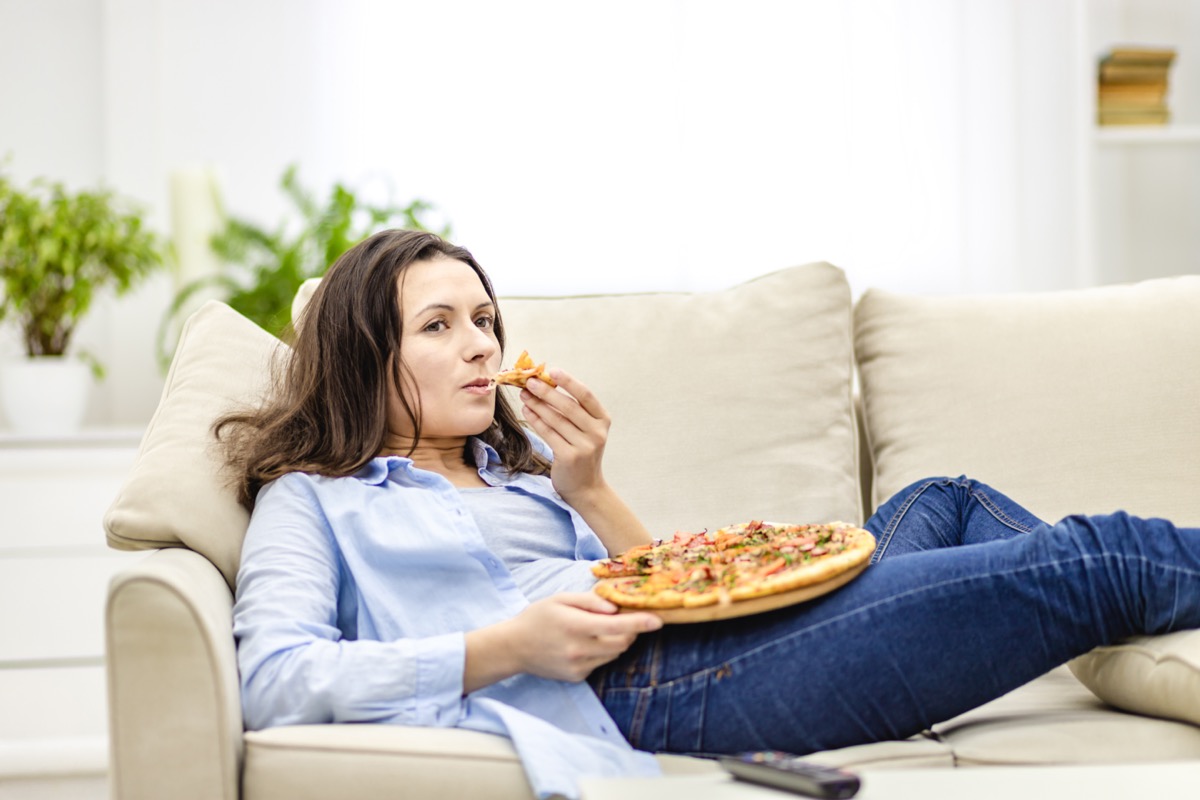 woman is chewing pizza, while laying on the white sofa. She is watching TV shows, being on blurred background.