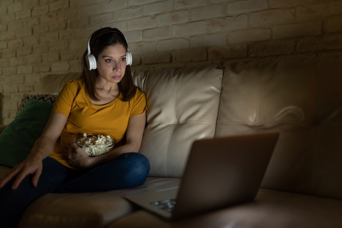 A woman in her 40s wearing headphones and eating popcorn while watching a movie on a streaming service on a laptop at night.