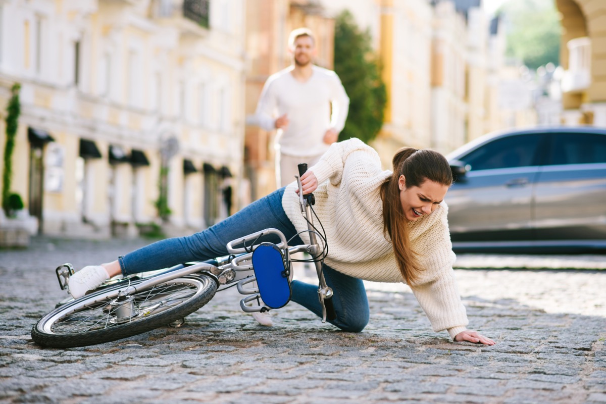 Woman on a city street falling with a bicycle on the road and a man running hurrying behind.
