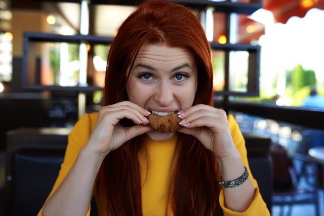 Woman biting crispy meat and being hungry, photo taken inside fast food restaurant, looking at camera with raised eyebrow.