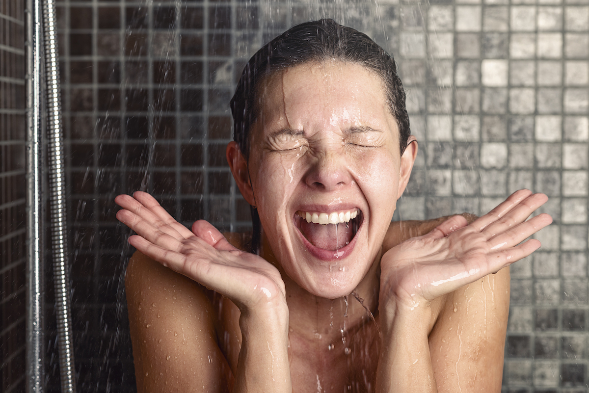 Young woman reacting in shock to hot or cold shower water as she stands under the shower head washing her hair eyes closed with her hands raised and mouth open
