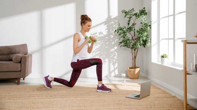 Tampilan samping wanita muda aktif dalam pakaian olahraga melakukan latihan lunges dengan dumbel dan menonton video di laptop selama latihan kebugaran di rumah