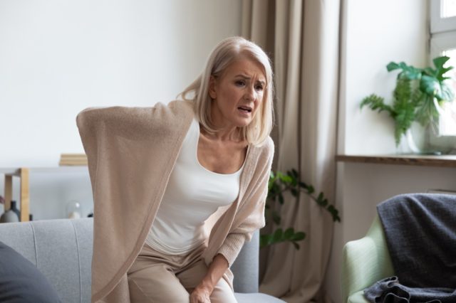 Middle aged mature woman suddenly feels hurt at home alone touching the sore spine.