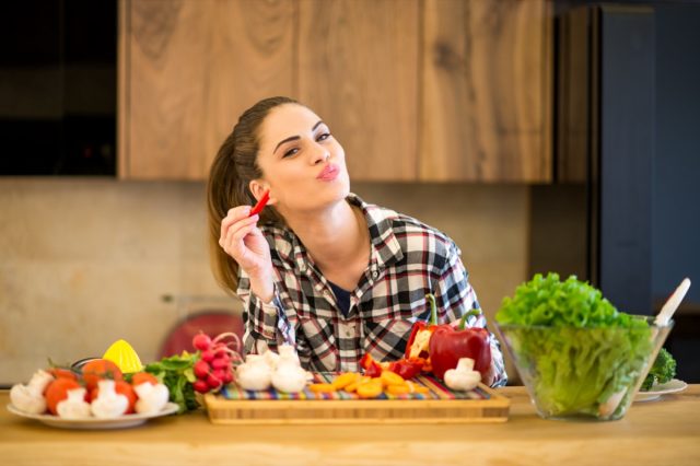 Young woman in the kitchen eating slice of red pepper.