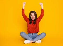 Full size photo of excited energetic girl sit floor legs crossed celebrate lottery win raise fists scream wear jeans jumper isolated bright shine color background