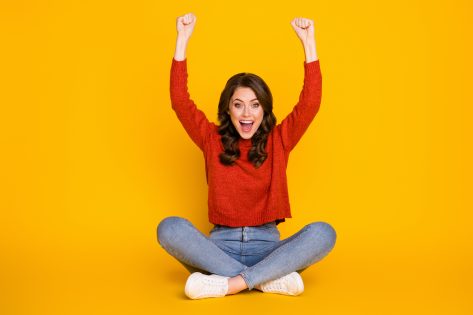 Full size photo of excited energetic girl sit floor legs crossed celebrate lottery win raise fists scream wear jeans jumper isolated bright shine color background