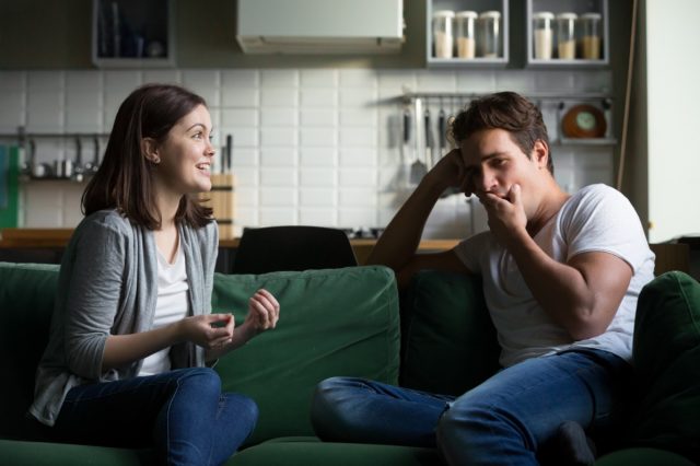 Man is yawning getting bored listening to excited woman talking while sitting on couch at home.