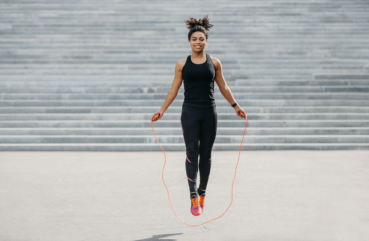 Female athlete does cardio workout. Smiling african american girl in sportswear with fitness tracker jumping rope outdoors, free space