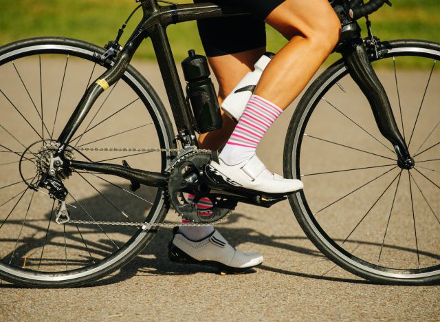 Legs of a female cyclist sitting on a pro slim bike. Cropped, no head. She's wearing striped white and pink socks and low top sneakers with velcro straps.