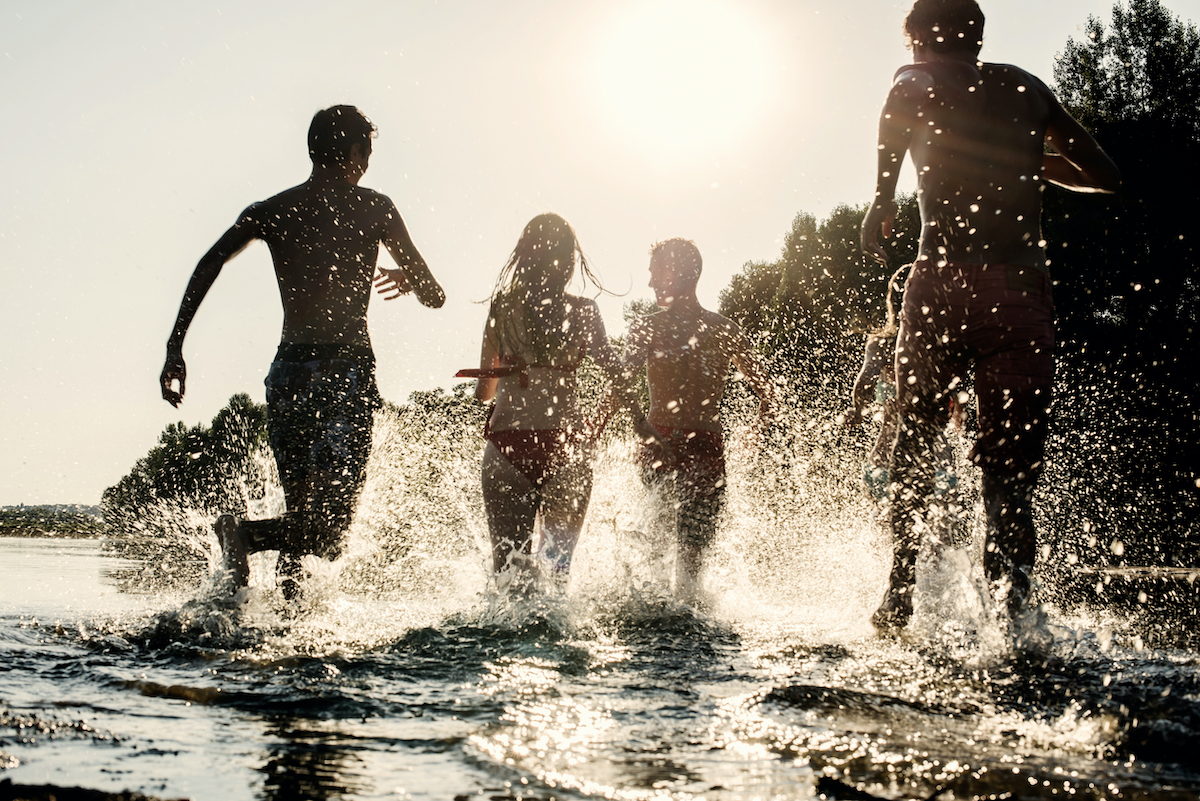 Backlit shot of teenagers running with their feet in the water at the beach during a sunny afternoon during summer break. They are having fun, playing and splashing water around them