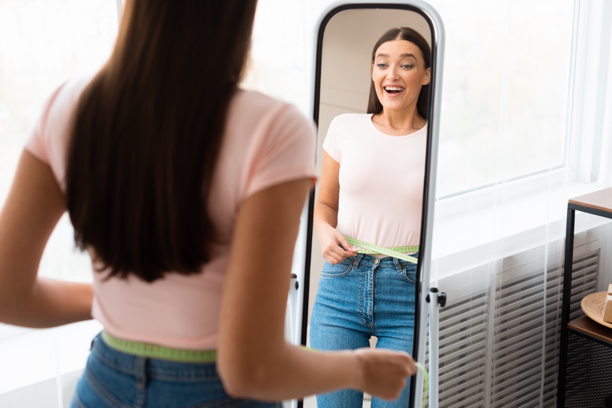 Woman measuring waist with tape standing in front of mirror.