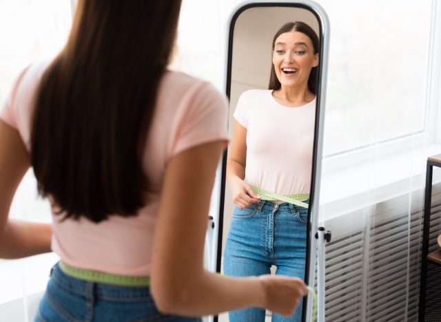 Woman measuring waist with tape standing in front of mirror.