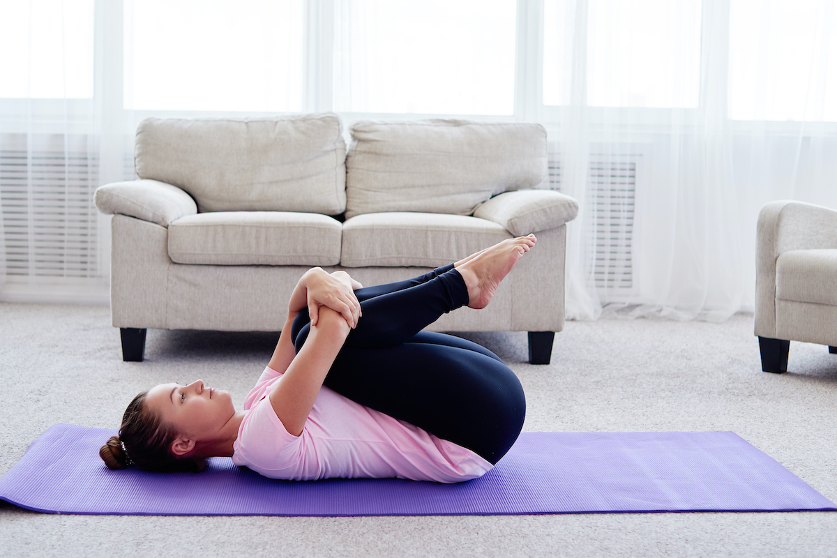 Portrait of young woman practicing apanasana pose on mat at home indoor, copy space. Apanasana. Knees to chest pose. Practicing yoga. Wellness and healthy lifestyle