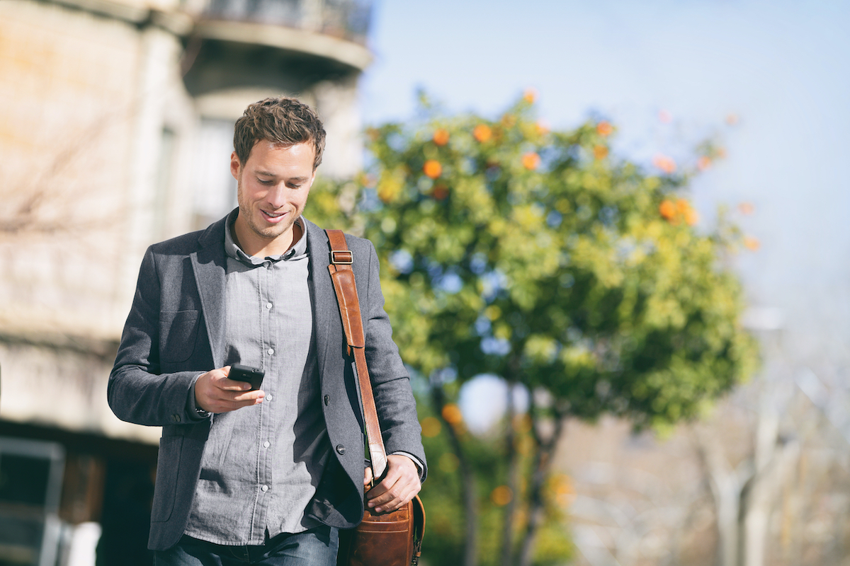 Business man using mobile phone walking in city street commuting to work with blazer and messenger bag texting on smartphone. Young businessman urban lifestyle.