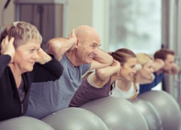 Happy elderly couple exercising in a pilates class at the gym with three other younger people toning and strengthening their muscles using gym balls, focus to the senior man and woman