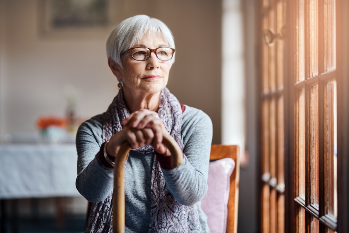 A senior woman holding cane looking through the window in a retirement home.