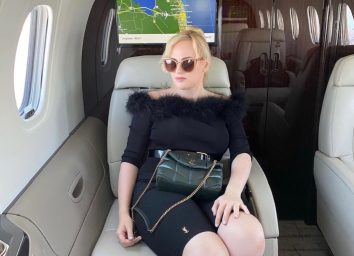 full body shot of rebel wilson sitting on plane in black outfit