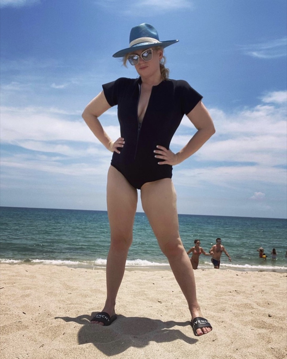 rebel wilson in black swimsuit and hat on the beach