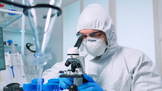 Ein Biotechnologe im PSA-Anzug sucht in einem Labor mit einem Mikroskop nach DNA.  Ein Team untersucht die Entwicklung des Virus mithilfe von Hochtechnologie für die wissenschaftliche Forschung, um einen Impfstoff gegen Covid19 zu entwickeln