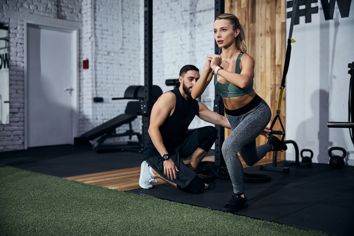 Person locking her hands together while doing a single-leg squat with an instructor placing her leg in a strap loop