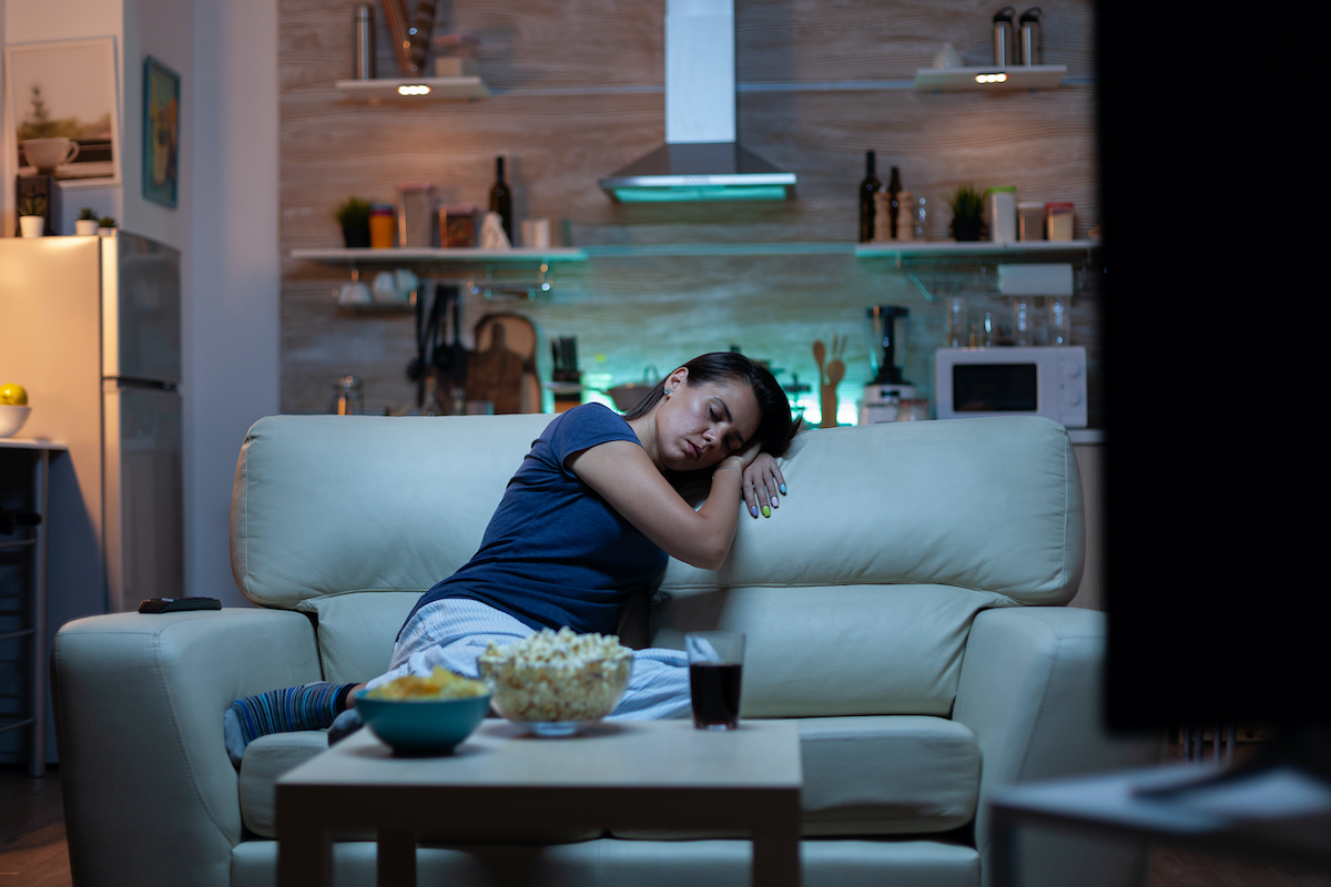 Woman falling asleep on sofa in front TV. Tired exhausted lonely sleepy lady in pajamas sleeping in front of television sitting on cozy couch in living room, closing eyes while watching movie at night