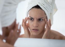 Unhappy woman wearing white bath towel checking skin after shower, looking in mirror, touching face skin.