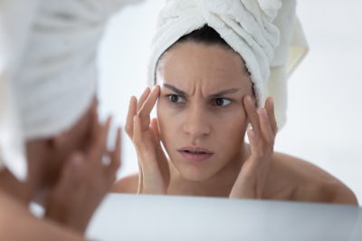 Unhappy woman wearing white bath towel checking skin after shower, looking in mirror, touching face skin.